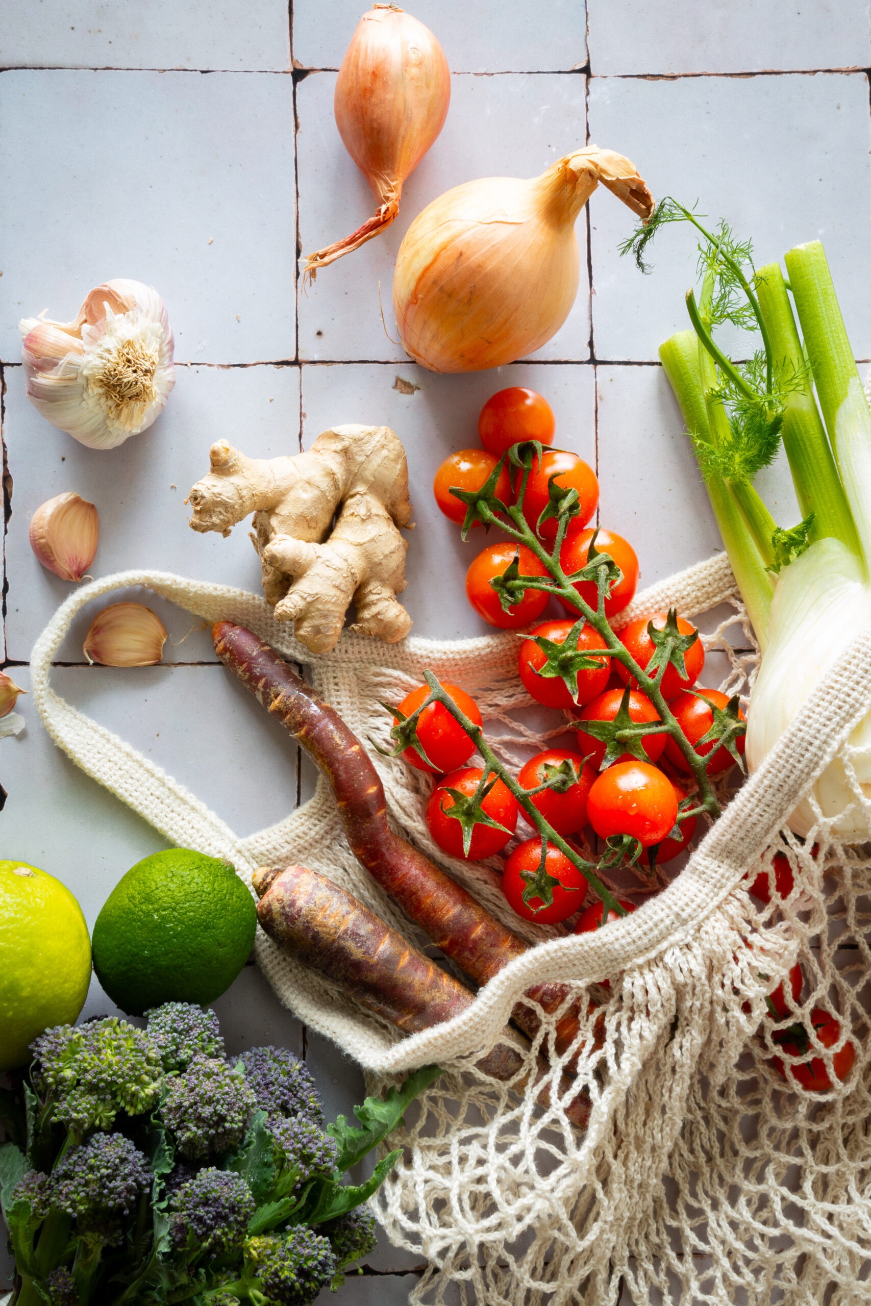 Gemüseeinkauf, der aus einer Stofftasche purzelt. Tomaten, Fenchel, Zwiebeln, Knoblauch, Ingwer, Limetten und Brokkoli.