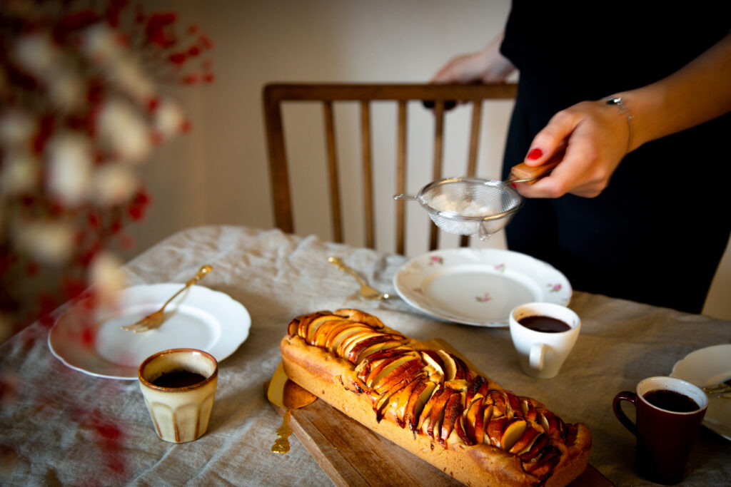 Gedeckter Tisch mit Apfelkuchen und Espresso. Der Kuchen wird im Bild gerade mit Puderzucker bestäubt.